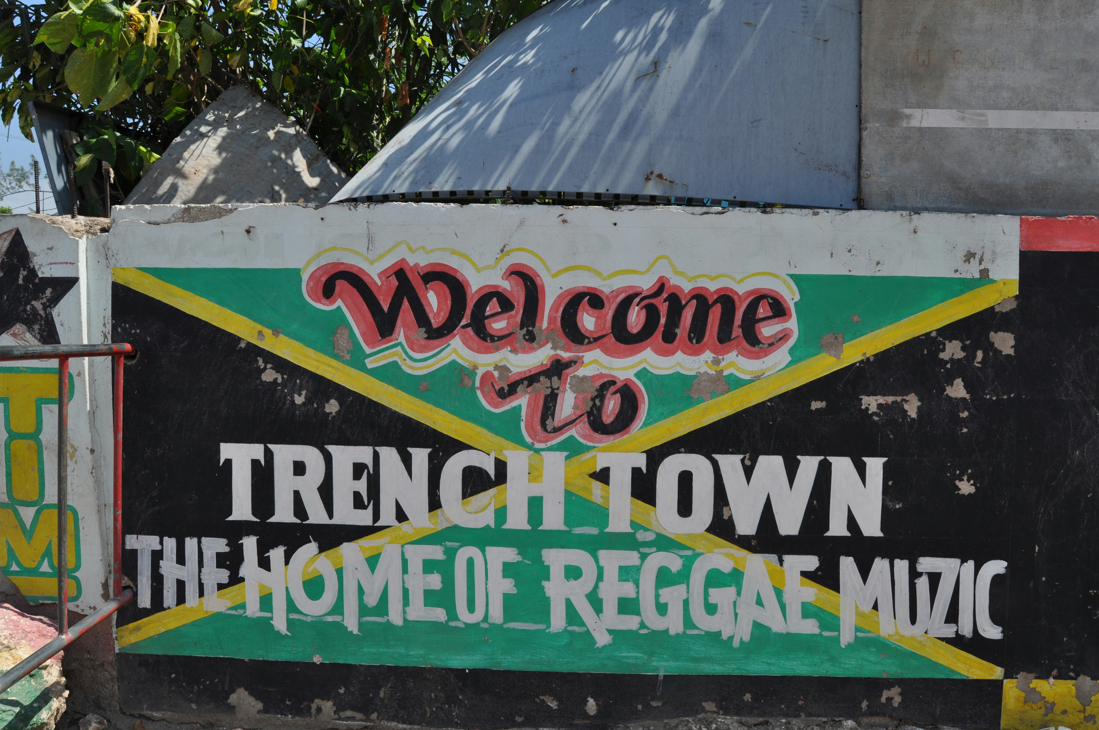 Concrete wall with the words "Trench Town The Home of Reggae Muzic" painted on top of a painting of the Jamaica green, black and gold flag.